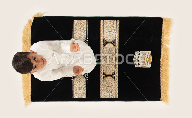 الدعاء والتقرب من الله تعالى, بورتريه علوي عامودي لطفل عربي خليجي سعودي مبتسم يرتدي الثوب التقليدي يقوم بالدعاء الى الله ينظر الى الكاميرا, مفهوم أركان الإسلام, الجلوس على سجادة الصلاة والقيام بالأذكار والأدعية اليومية, إسلاميات وعبادة, خلفية بيضاء