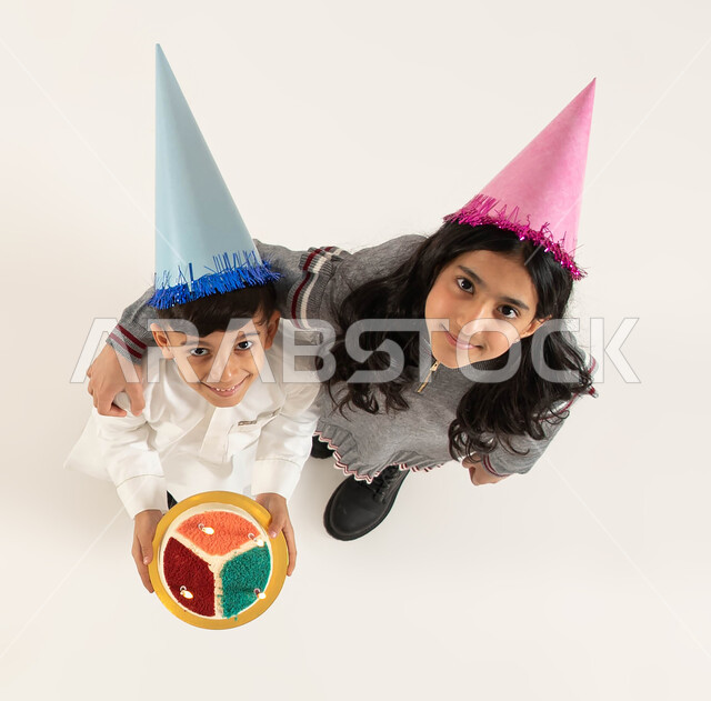 A boy wearing a traditional costume holding a cake, gestures of happiness and pleasure, spending fun times and enjoying birthdays and family events, a vertical overhead portrait of two smiling Saudi Arabian Gulf brothers wearing a birthday hat looking at 