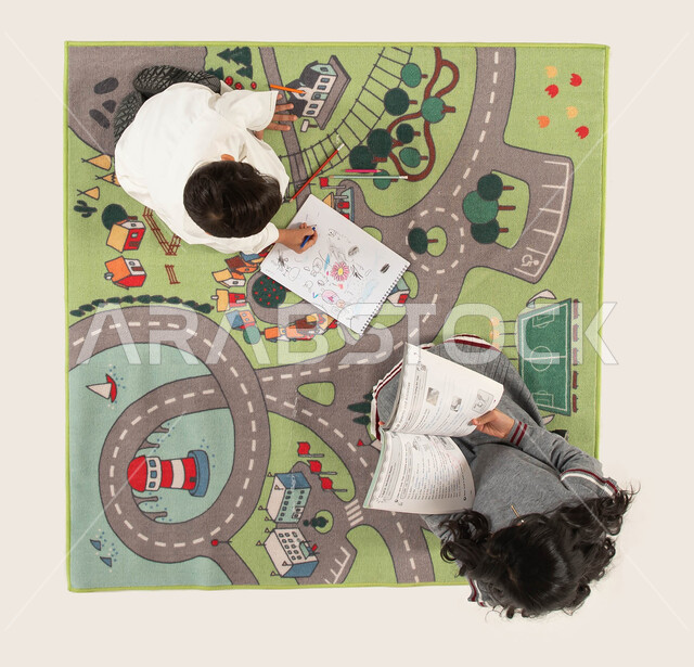 الاستمتاع بالمطالعة وقراءة الكتب المفيدة, قضاء أوقات ممتعة مع الأخوات, تقوية الإبداع الفني لدى الأبناء واستخدام أدوات الرسم والتلوين, صورة علوية عامودية لأخوين عربيين سعوديين خليجيين يجلسان على سجادة عصرية في غرفة الاطفال.