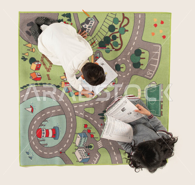 تقوية الإبداع الفني لدى الأبناء واستخدام أدوات الرسم والتلوين, صورة علوية عامودية لأخوين عربيين سعوديين خليجيين يجلسان على سجادة عصرية في غرفة الاطفال، قضاء أوقات ممتعة مع الأخوات, الاستمتاع بالمطالعة وقراءة الكتب المفيدة.