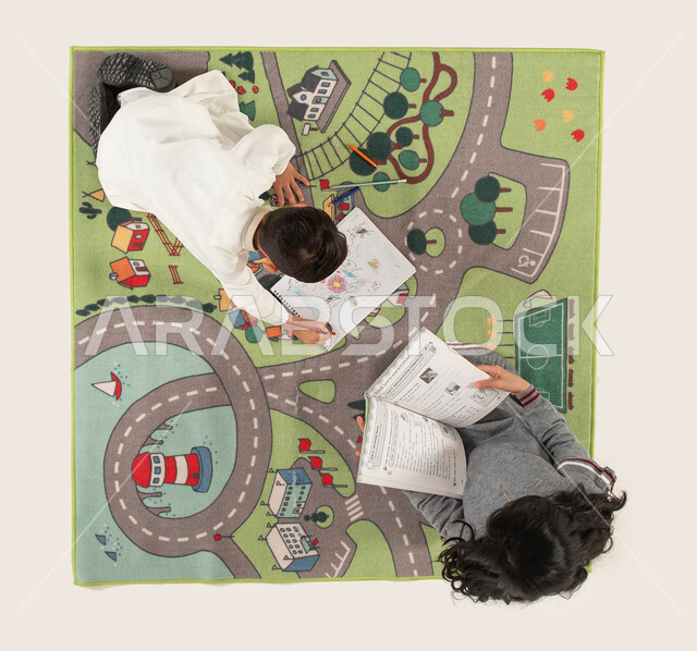 قضاء أوقات ممتعة مع الأخوات, الاستمتاع بالمطالعة وقراءة الكتب المفيدة, تقوية الإبداع الفني لدى الأبناء واستخدام أدوات الرسم والتلوين, صورة علوية عامودية لأخوين عربيين سعوديين خليجيين يجلسان على سجادة عصرية في غرفة الاطفال.