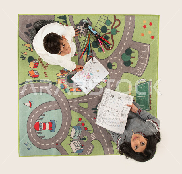 تنيمة المهارات الفنية لدى الأبناء واستخدام أدوات الرسم والتلوين, صورة علوية عامودية لأخوين عربيين سعوديين خليجيين مبتسمان  يجلسان على سجادة عصرية في غرفة الاطفال ينظران الى الكاميرا، قضاء أوقات ممتعة مع الأخوات, الاستمتاع بالمطالعة وقراءة الكتب المفيدة.