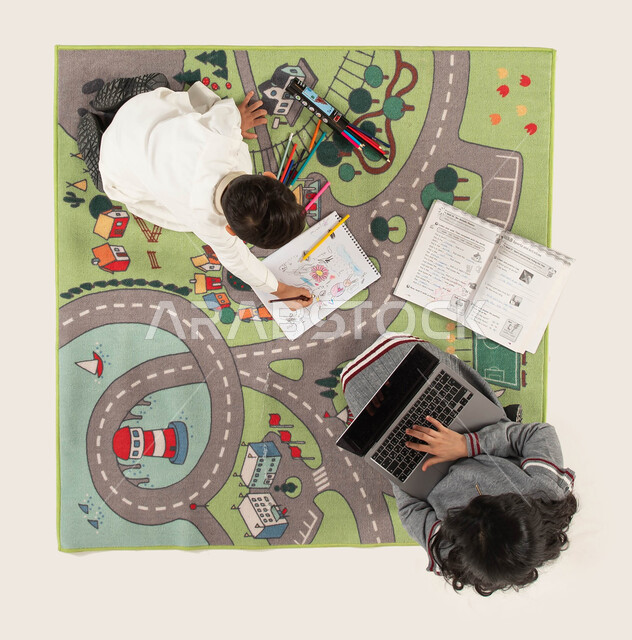 الاستمتاع بممارسة الهوايات المختلفة في أوقات الفراغ, صورة عامودية علوية لأخوين عربيين سعوديين خليجيين يجلسان على سجادة عصرية بغرفة الأولاد, البحث عن معلومة ما على الانترنت, استخدام أدوات الرسم والتلوين وتقوية الإبداع الفني لدى الأبناء.