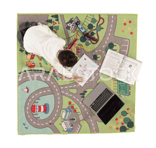 استخدام أدوات الرسم والتلوين وتقوية الإبداع الفني لدى الأبناء، انشطة ترفيهية ثقافية ممتعة للاطفال، صورة علوية عامودية لولد عربي خليجي سعودي يرتدي الزي التقليدي يجلس على سجاد بتصميم عصري يقوم بحل الواجب، دمج العملية التعليمية مع التقنية والتكنولوجيا.