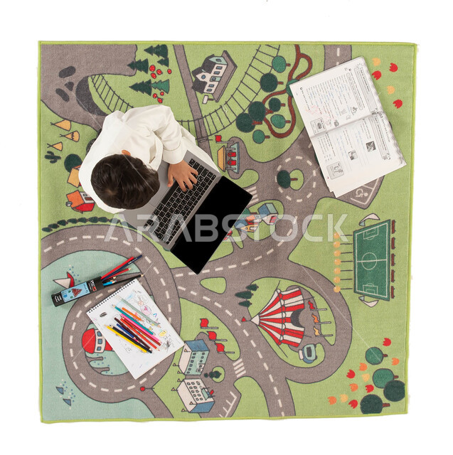 دورات اونلاين تعلمية ثقافية وانشطة ترفيهية للاطفال، التعلم وتلقي الدروس عن بعد، صورة علوية عامودية لولد عربي خليجي سعودي يرتدي الزي التقليدي يجلس على سجاد بتصميم عصري يستخدم جهاز الحاسوب المحمول، دمج العملية التعليمية مع التقنية و التكنولوجيا.