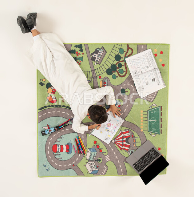 انشطة ترفيهية ثقافية ممتعة للاطفال، استخدام أدوات الرسم والتلوين وتقوية الإبداع الفني لدى الأبناء، صورة علوية عامودية لولد عربي خليجي سعودي يرتدي الزي التقليدي يجلس على سجاد بتصميم عصري يقوم بحل الواجب، دمج العملية التعليمية مع التقنية والتكنولوجيا.