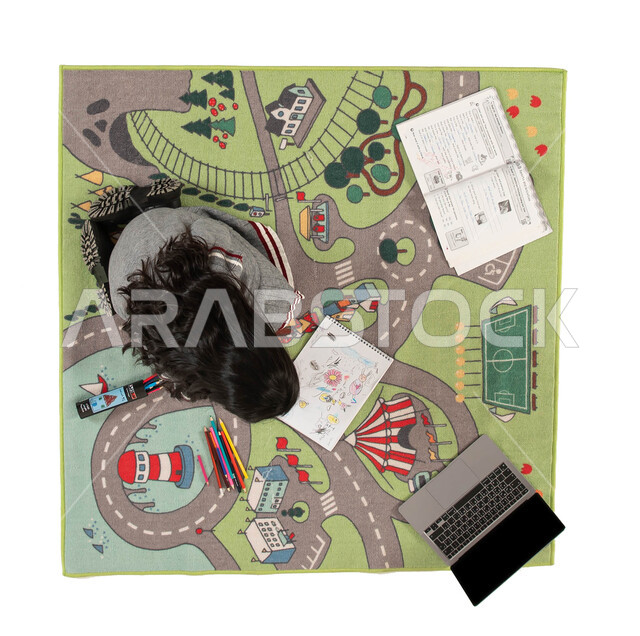 تنمية المواهب الفنية، انشطة ترفيهية ودورات اونلاين تعلمية ثقافية للاطفال، التعلم وتلقي الدروس عن بعد، صورة علوية عامودية لفتاة عربية خليجية سعودية تجلس على سجاد بتصميم عصري تقوم بالرسم والتلوين وحل الواجبات، دمج العملية التعليمية مع التقنية و التكنولوجيا.