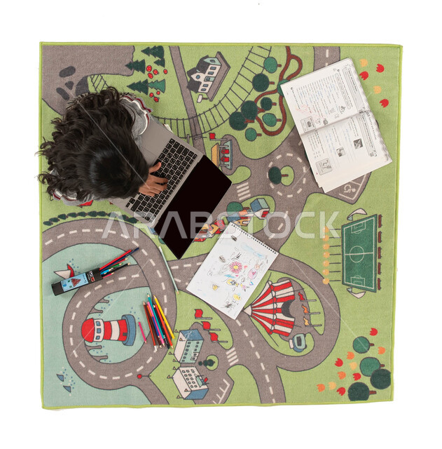 Online courses, educational, cultural and entertainment activities for children, learning and taking lessons from a distance, a vertical overhead image of a Saudi Gulf Arab girl sitting on a carpet with a modern design using a laptop, integrating the educ