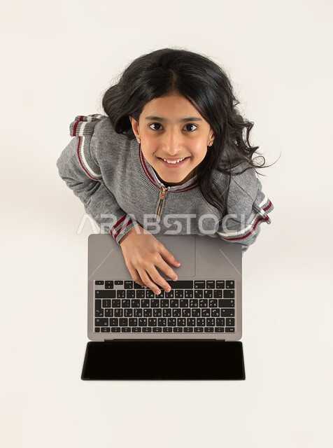 دورات تعلمية ثقافية و انشطة ترفيهية الكترونية للاطفال، التعلم وتلقي دروس الاونلاين عن بعد، بورتريه علوي عامودي لفتاة عربية خليجية سعودية مبتسمة تستخدم جهاز الحاسوب تنظر الى الكاميرا، قضاء اوقات مسلية باللعب في الحاسب المحمول، خلفيه بيضاء.