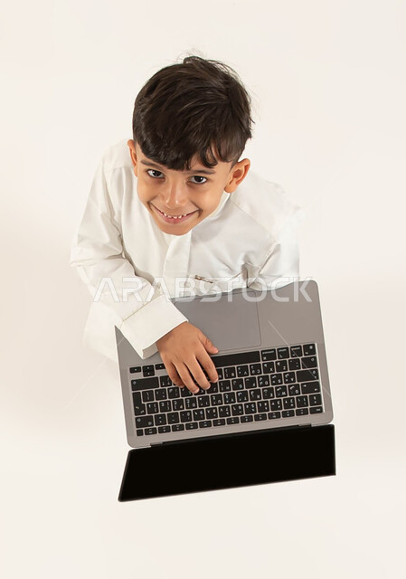 دورات تعلمية ثقافية و انشطة ترفيهية الكترونية للاطفال، التعلم وتلقي دروس الاونلاين عن بعد، بورتريه علوي عامودي لفتى عربي خليجي سعودي مبتسم يرتدي الزي التقليدي يحمل جهاز الحاسوب ينظر الى الكاميرا، قضاء اوقات مسلية باللعب في الحاسب المحمول، خلفيه بيضاء.