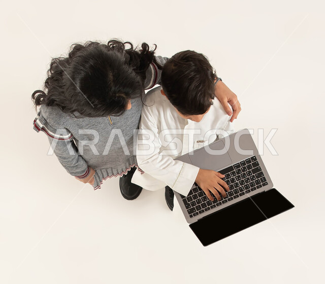  انشطة ترفيهيه الكترونية مسلية للاطفال، بورتريه علوي عامودي لولد وبنت عربيان خلجيان سعوديان يقومان بإستخدام الحاسوب، تلقي دروس الاونلاين والتعلم عن بعد عبر الحاسب المحمول، قضاء اوقات ممتعة برفقة الاخوة، خلفية بيضاء