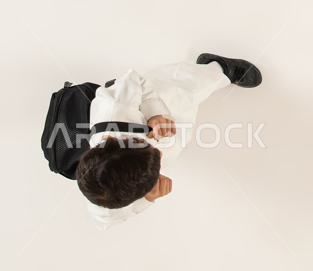 التحضير لبداية عام دراسي جديد، الاستعداد والتجهز للذهاب الى المدرسة، بورتريه علوي عامودي لطالب عربي خليجي سعودي يرتدي الزي التقليدي يحمل حقيبه الظهر المدرسيه، ايماءات الحماس و النشاط بالعودة الى الدراسة، التعليم في المملكه العربية السعوديه، خلفيه بيضاء.