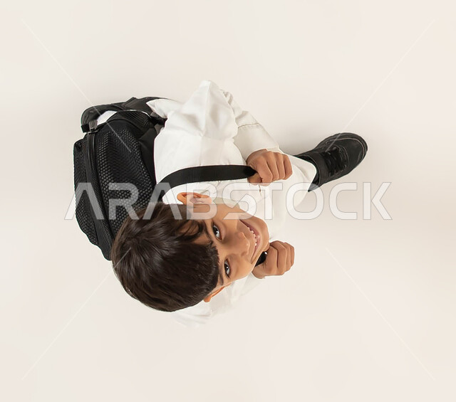 الاستعداد والتجهز للذهاب الى المدرسة، بورتريه علوي عامودي لطالب عربي خليجي سعودي مبتسم  يرتدي الزي التقليدي يحمل حقيبه الظهر المدرسيه ينظر الى الكاميرا، التحضير لبداية عام دراسي جديد، ايماءات السعادة و السرور للعودة الى الدراسة، خلفيه بيضاء.