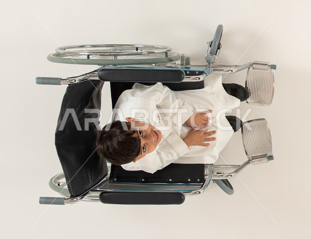 تأمين اجهزة ومعدات تساعد بالحركة والتنقل، مريض يجلس على كرسي متحرك، ايماءات التفاؤل بالحياة المستقبلية، تحسين حياة أصحاب الهمم وتأمين احتياجاتهم الخاصة، بورتريه علوي عامودي لولد عربي خليجي سعودي من ذوي الاحتياجات الخاصه ينظر الى الكاميرا، خلفيه بيضاء