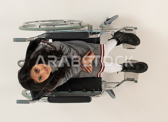 ايماءات التفاؤل بالحياة المستقبلية، مريضه تجلس على كرسي متحرك، تحسين حياة أصحاب الهمم وتأمين احتياجاتهم الخاصة، بورتريه علوي عامودي لفتاة عربيه خليجيه سعودية من ذوي الاحتياجات الخاصه تنظر الى الكاميرا، تأمين اجهزة ومعدات تساعد بالحركة والتنقل، خلفيه بيضاء