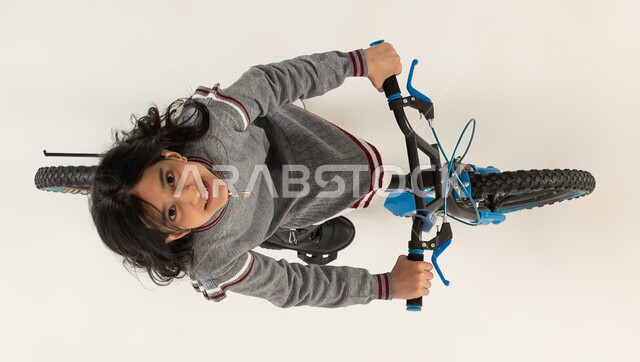 ممارسة هواية ركوب الدراجات الهوائيه، بورتريه علوي عامودي لفتاة عربية خليجية سعودية مبتسمة  تقود الدراجه  تنظر الى الكاميرا، قضاء الأوقات في تعلم أنشطة ترفيهية ممتعة للأطفال، نشاط بدني للحفاظ على اللياقة الجسدية، المشاركة في سباق الدراجات، خلفيه بيضاء.