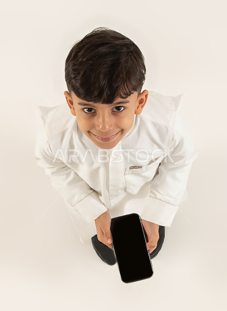 عرض برامج وتطبيقات الهاتف المحمول، بورتريه علوي عامودي لولد عربي خليجي سعودي مبتسم يرتدي الزي التقليدي يحمل بيده هاتف محمول بشاشه سوداء، إستخدام مواقع ووسائل التواصل الاجتماعي، مفهوم الإعلانات والتسويق الالكتروني، خلفيه بيضاء.