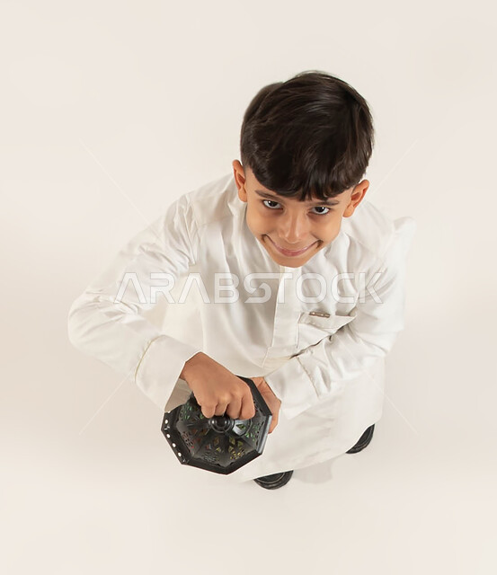 التزيين والتحضير للأجواء الروحانيه الرمضانيه، الأعياد الإسلامية والمناسبات الدينيه، التجهيزات لاستقبال شهر رمضان المبارك، بورتريه علوي عامودي لصبي عربي خليجي سعودي مبتسم يحمل بيده الفانوس الرمضاني ينظر الى الكاميرا، اسلاميات و عبادة، خلفيه بيضاء.