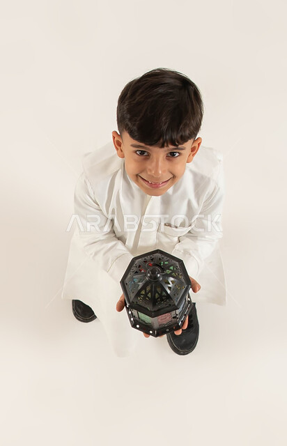 الأعياد الإسلامية والمناسبات الدينيه، التجهيزات لاستقبال شهر رمضان المبارك، بورتريه علوي عامودي لصبي عربي خليجي سعودي مبتسم يحمل بيده الفانوس الرمضاني ينظر الى الكاميرا، التزيين والتحضير للأجواء الروحانيه الرمضانيه، اسلاميات و عبادة، خلفيه بيضاء.