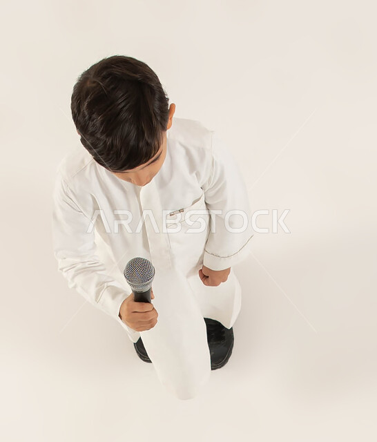 اكتشاف مواهب الأطفال وتنميتها، بورتريه علوي عامودي لصبي عربي خليجي سعودي يقوم بالغناء عبر المايكروفون، إيماءات الاندماج و التفاعل مع الموسيقى، تقديم العروض الفنيه الغنائيه والفقرات الترفيهية المسليه، تسجيل مقطع صوتي غنائي، خلفيه بيضاء.