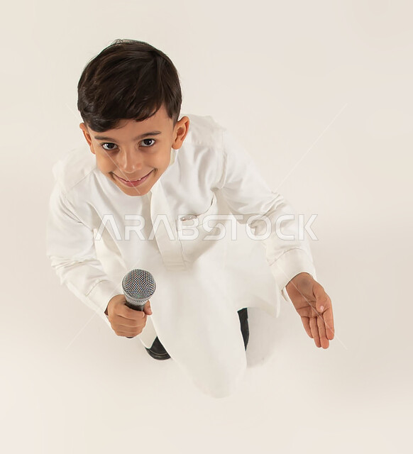 موهبة الغناء والتدريب الصوتي، بورتريه علوي عامودي لصبي عربي خليجي سعودي مبتسم يمسك بيده المايكروفون ينظر إلى الكاميرا، تقديم العروض الفنيه المسرحيه الغنائيه والفقرات الترفيهية المسليه، إيماءات تدل على الحماس والسعاده، تنميه مواهب الأطفال، خلفيه بيضاء.