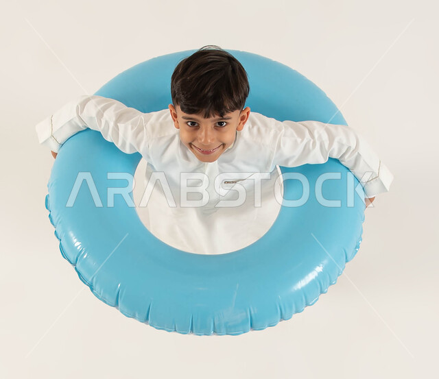 الاستعداد للذهاب في رحلة بحرية، بورتريه علوي عامودي لولد عربي خليجي سعودي مبتسم يقف بداخل عوامه السباحه الزرقاء ينظر إلى الكاميرا، الإستمتاع بعطله صيفيه سعيده، ايماءات الحماس والفرح لبداية التدرب على السباحة، هوايات ومهارات رياضيه بدنية، خلفيه بيضاء.