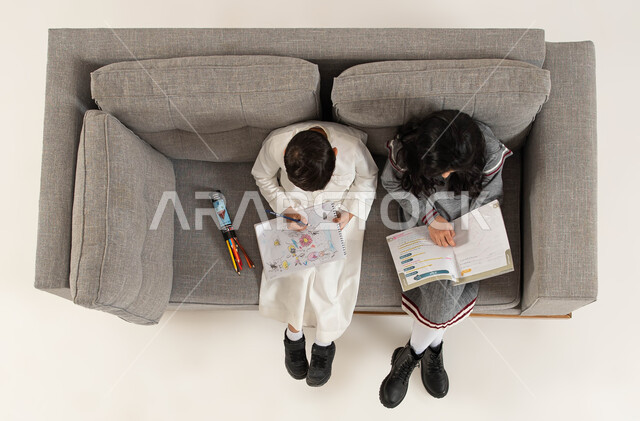 التعليم وتنمية المهارات الفنية والذهنية، بورتريه علوي عامودي لأخوين عربيين خليجيين سعوديين يجلسان على الأريكه يقومان باستغلال أوقات الفراغ بالأنشطة التعليمية الثقافية، المطالعة وقراءة الكتب المفيده، قضاء وقت ممتع بالرسم والتلوين، خلفية بيضاء