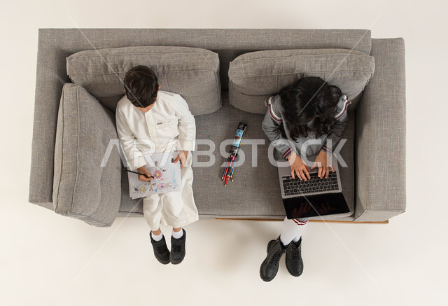 تلقي دروس الاونلاين والتعلم عن بعد عبر الحاسب المحمول، بورتريه علوي عامودي لأخوين عربيين خليجيين سعوديين يجلسان على الأريكه يقومان بإستغلال أوقات الفراغ بالأنشطه الالكترونية والواقعيه المفيده، قضاء وقت ممتع في تعلم الرسم والتلوين، خلفيه بيضاء.