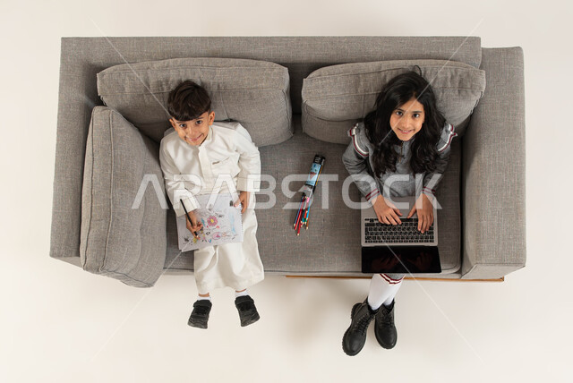 التعلم عن بعد وتلقي دروس الاونلاين عبر الحاسب المحمول، بورتريه علوي عامودي لأخوين عربيين خليجيين سعوديين يجلسان على الأريكه ينظران الى الكاميرا، قضاء وقت ممتع في تعلم الرسم والتلوين، إستغلال أوقات الفراغ بالأنشطه الالكترونية والواقعيه المفيده، خلفيه بيضاء