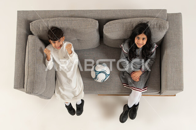 الاستمتاع بمشاهدة المباريات وبطوله كأس العالم، ايماءات تدل على الفرح بالفوز والإنتصار في مباراة كرة القدم، قضاء أوقات ممتعة برفقة الأصدقاء، بورتريه علوي عامودي لولد وبنت عربيان خليجيان سعوديان  يجلسان على الاريكة ينظران الى الكاميرا، خلفيه بيضاء.