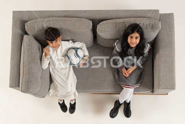 ايماءات تدل على الفرح بالفوز والإنتصار، بورتريه علوي عامودي لأخوان عربيان خليجيان سعوديان مبتسمان يجلسان على الاريكة ينظران الى الكاميرا، الشغف بمباريات كره القدم وبطوله كأس العالم، قضاء أوقات ممتعة برفقة الأصدقاء، مشاهدة مباراة كرة القدم، خلفيه بيضاء.