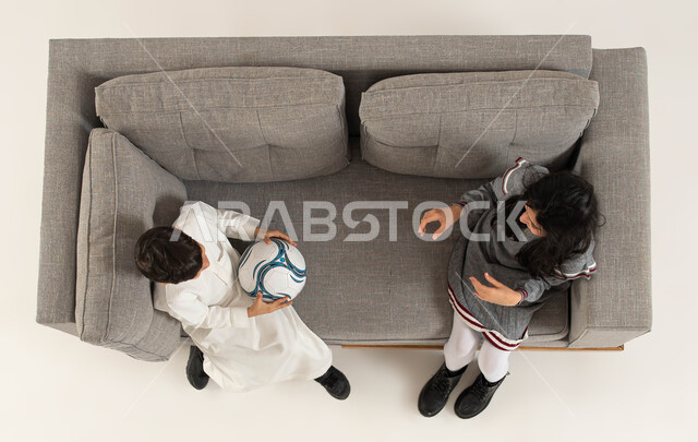  قضاء أوقات ممتعة برفقة الأصدقاء، بورتريه علوي عامودي لولد وبنت عربيان خليجيان سعوديان يجلسان على الاريكة ويقومان باللعب في الكرة، الشغف بالمبارايات و الرياضات منذ الصغر، تحدي مباراة كرة القدم، خلفيه بيضاء.