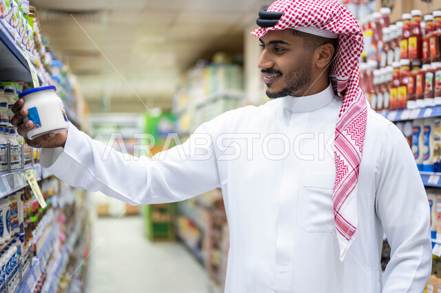 الاستفادة من العروض والتخفيضات على المنتجات, الاستمتاع بقضاء وقت ممتع في التسوق بالمحلات التجارية, عرض منتجات وتموينات محلية ومستوردة في السوبرماركت, رجل عربي خليجي سعودي مبتسم يرتدي الثوب التقليدي والشماغ يقوم باختيار أفضل أنواع الجبن الكريمي
