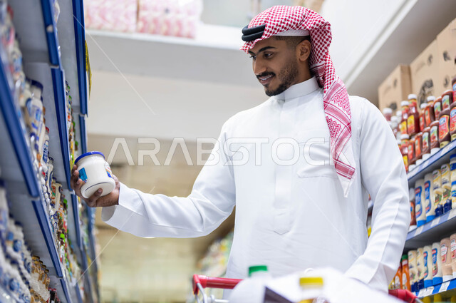 الاستمتاع بقضاء وقت ممتع في المحلات التجارية, عرض منتجات وتموينات محلية ومستوردة في السوبرماركت, رجل عربي خليجي سعودي مبتسم يرتدي الثوب التقليدي والشماغ يقوم باختيار  أفضل أنواع الجبن الكريمي, الاستفادة من العروض والتخفيضات على مستلزمات المستهلك السعودي