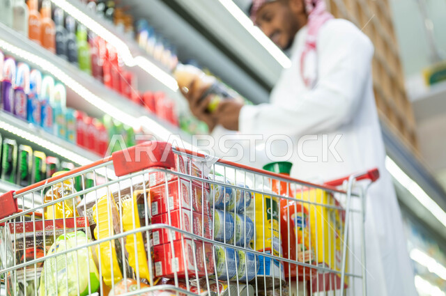 أصناف وأنواع من المنتجات ومستلزمات المستهلك من سلع ومواد غذائيه مختلفه في المراكز التجاريه, صورة مقربة لسلة التسوق المليئة بالأغراض واللوازم المنزلية, رجل عربي سعودي خليجي يقوم بالتبضع والتسوق واختيار الأغذية الوطنية من الأسواق السعودية