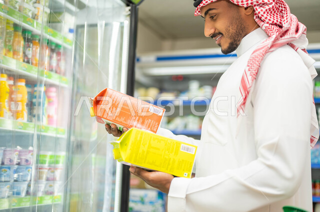 الاستفادة من العروض والتخفيضات على مستلزمات المستهلك السعودي, عرض منتجات وتموينات محلية في الماركت, صورة من الجانب لرجل عربي خليجي سعودي مبتسم يرتدي الثوب التقليدي والشماغ يقف في قسم العصائر والمشروبات, شراء وتسوق واختيار المستلزمات الغذائية المنزلية