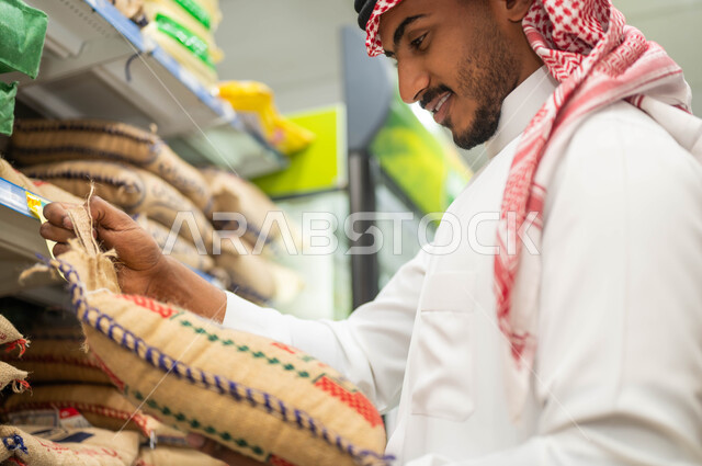 مجموعة من أكياس الأرز على الرفوف داخل السوبرماركت, صورة من الجانب لرجل عربي خليجي سعودي مبتسم يرتدي الثوب التقليدي يقوم باختيار الأرز المناسب للطبخ السعودي, محاصيل زراعية وتموينات محلية في الماركت، الاستفادة من العروض والتخفيضات على المنتجات السعودية