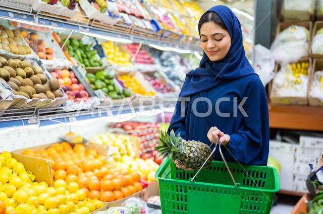 محاصيل زراعية ومنتجات وطنية محلية في البقالات السعودية, انتقاء أفضل أنواع الفاكهة والقيام بالتسوق وشراء احتياجات المستهلك السعودي من المراكز التجاريه، صوره مقربه لسيدة تحمل سلة التسوق مبتسمة محجبة تقوم بحمل الأناناس في قسم الفواكه والخضار