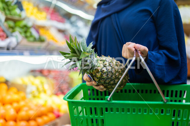 Shopping and purchasing the needs of the Saudi consumer from one of the commercial centers, a close-up of the hand of a woman carrying a shopping basket wearing an abaya carrying pineapples in the fruits and vegetables section, selecting the best types of