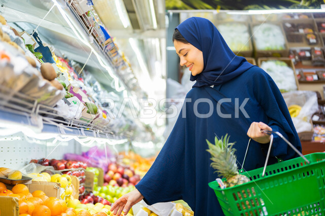 Doing shopping and purchasing the needs of the Saudi consumer from one of the commercial centers and benefiting from offers and discounts. A close-up of a smiling veiled young woman carrying a shopping basket and picking oranges in the fruits and vegetabl