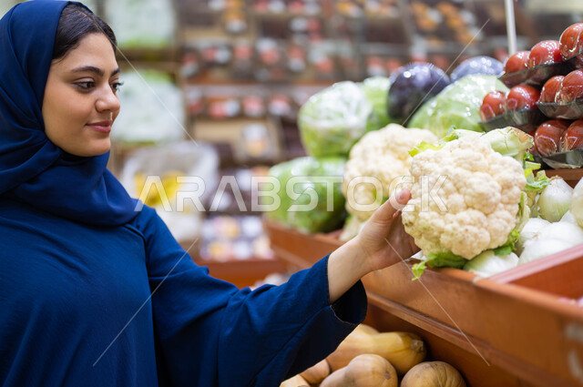 الاستمتاع بقضاء وقت ممتع في التسوق, التجول في إحدى المحلات التجارية في السعودية, صورة مقربة لسيدة عربية خليجية مبتسمة محجبة ترتدي العباءة تقوم باختيار القرنبيط من قسم الخضار والفواكه, محاصيل زراعية ومنتجات محلية ومستوردة في الماركت