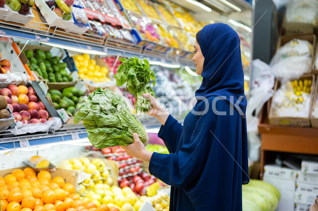 تأمين الاحتياجات المنزلية الغذائية الصحية, التبضع في قسم الخضراوات واختيار الأجود والأفضل من المحلات التجارية في السعودية, صورة من الجانب لشابة عربية سعودية خليجية مبتسمة محجبة ترتدي العباءة وتقوم بشراء مستلزمات ومكونات الطبخ والغذاء الصحي