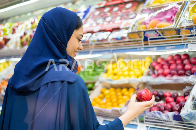شراء الاحتياجات المنزلية من قسم الخضراوات والفواكه في المحلات التجارية, صورة مقربة من الخلف لشابة عربية سعودية خليجية محجبة مبتسمة تمسك فاكهه الخوخ الطازجة, قضاء وقت ممتع في التسوق واختيار السناكات والأغذية الصحية, محاصيل زراعية وطنية محلية في البقالة