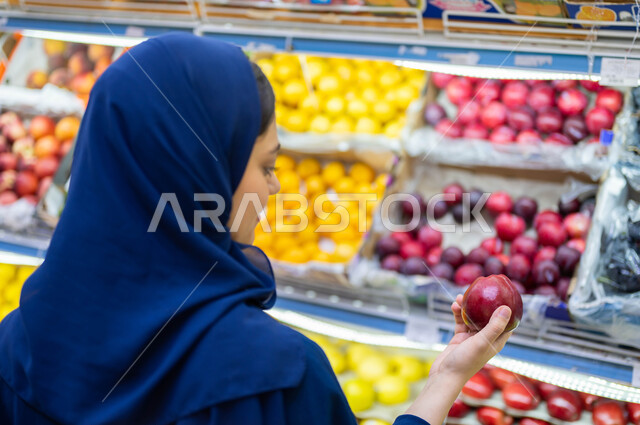 شراء الاحتياجات المنزلية من قسم الخضراوات والفواكه في المحلات التجارية, صورة مقربة من الخلف لشابة عربية سعودية خليجية محجبة تمسك فاكهه الخوخ الطازجة, قضاء وقت ممتع في التسوق بالأسواق السعودية واختيار السناكات والأغذية الصحية