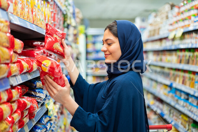 Preparing to buy supplies from Saudi commercial centers, taking advantage of discounts and offers on products, a side image of a Saudi Arabian Gulf woman wearing a black abaya, choosing the best types of pasta from the shelves of the supermarket and spend