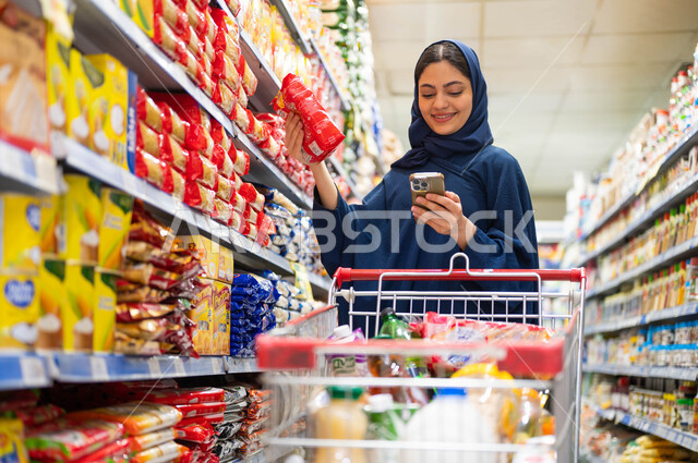 انتقاء أفضل أنواع المعكرونة من البقالة, مركز تجاري لتلبية احتياجات المستهلك السعودي, استخدام الهاتف المحمول في الماركت, السلع والمواد اللازمة للمتطلبات المنزلية, صورة مقربة لإمرأة عربية سعودية خليجية محجبة مبتسمة تقوم بقراءة قائمة الاحتياجات من الجوال