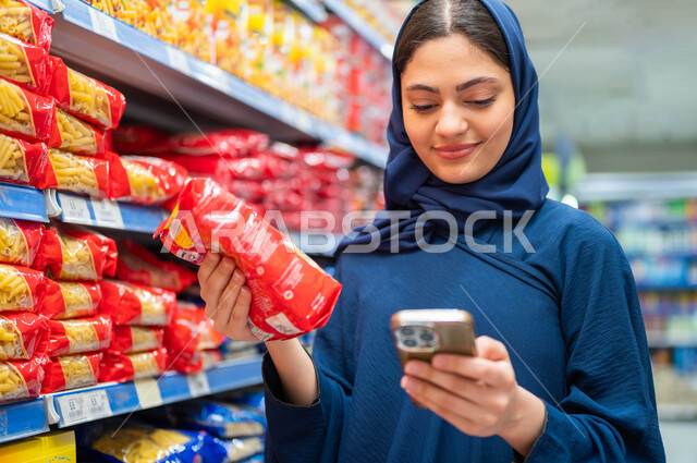 مركز تجاري لتلبية احتياجات المستهلك السعودي, استخدام الهاتف المحمول في الماركت, السلع والمواد اللازمة للمتطلبات المنزلية, صورة مقربة لإمرأة عربية سعودية خليجية محجبة مبتسمة تقوم بقراءة قائمة الاحتياجات من الجوال, انتقاء أفضل أنواع المعكرونة من البقالة