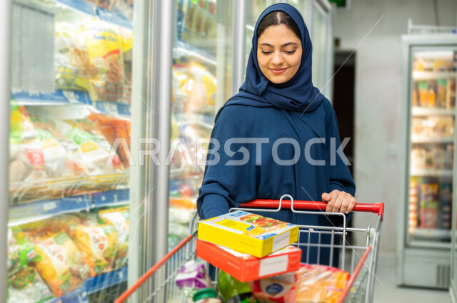 الاستمتاع بالتسوق في المتاجر والمحلات التجارية, السلع والمواد الغذائية المختلفة, إمرأة سعودية عربية خليجية تجر عربة التسوق المليئة بالأطعمة والمشروبات وتقوم بشراء الاحتياجات المنزلية من السوبرماركت, مستلزمات المستهلك السعودي
