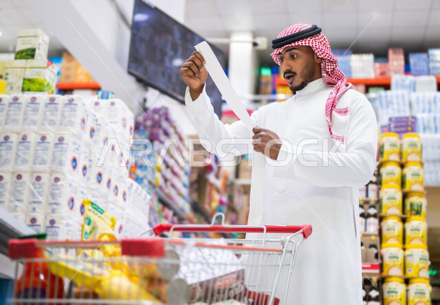 إصدار الفواتير عبر جهاز الحاسوب, قراءة قائمة المشتريات بإيماءات الدهشة والذهول, رجل عربي سعودي خليجي يرتدي الثوب التقليدي يقف في المتجر الغذائي, غلاء أسعار المستلزمات المنزلية, الاستفادة من العروض والتخفيضات على المنتجات الغذائية