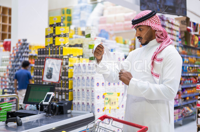 النظر إلى فاتورة المشتريات بإيماءات الدهشة والذهول, رجل عربي سعودي خليجي يرتدي الثوب التقليدي يقف عند الكاشير في السوبرماركت, غلاء أسعار المستلزمات المنزلية, إصدار الفواتير عبر جهاز الحاسوب, الاستفادة من العروض والتخفيضات على المنتجات الغذائية
